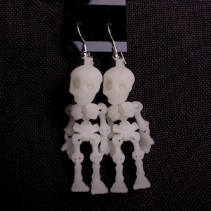 May include: A pair of white skeleton earrings. The earrings are made of plastic and are shaped like skeletons. The skeletons are wearing a simple, white dress.