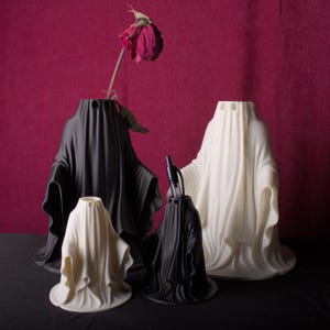 May include: A collection of ghost-shaped vases in black and white, with a dried rose in one. The vases vary in size and have a draped fabric design. The background is a deep red, creating a contrast with the vases.