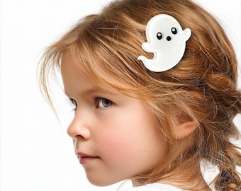 May include: A white ghost-shaped hair clip with black eyes and a small mouth is clipped into the hair of a young person. The hair is light brown and styled in a braid. The background is white.
