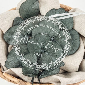 Könnte beinhalten: Ein transparenter Weihnachtsbaumschmuck aus Acryl mit einem weißen floralen Kranzdesign und dem Text "Erstes Weihnachten als Mama & Papa 2023".