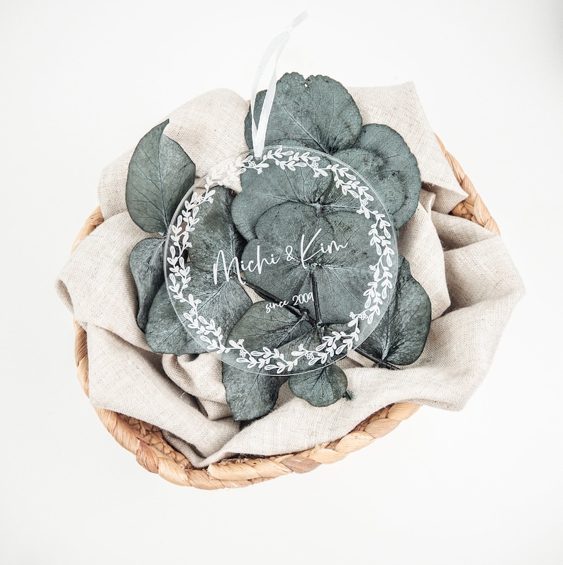 May include: A clear round ornament with a white floral wreath design and the text "Michi & Kim since 2004" on a beige fabric background.