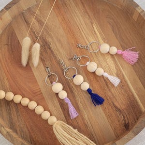 May include: Five wooden bead keychains with different colored tassels. The keychains are on a wooden surface. The tassels are pink, white, blue, and purple.