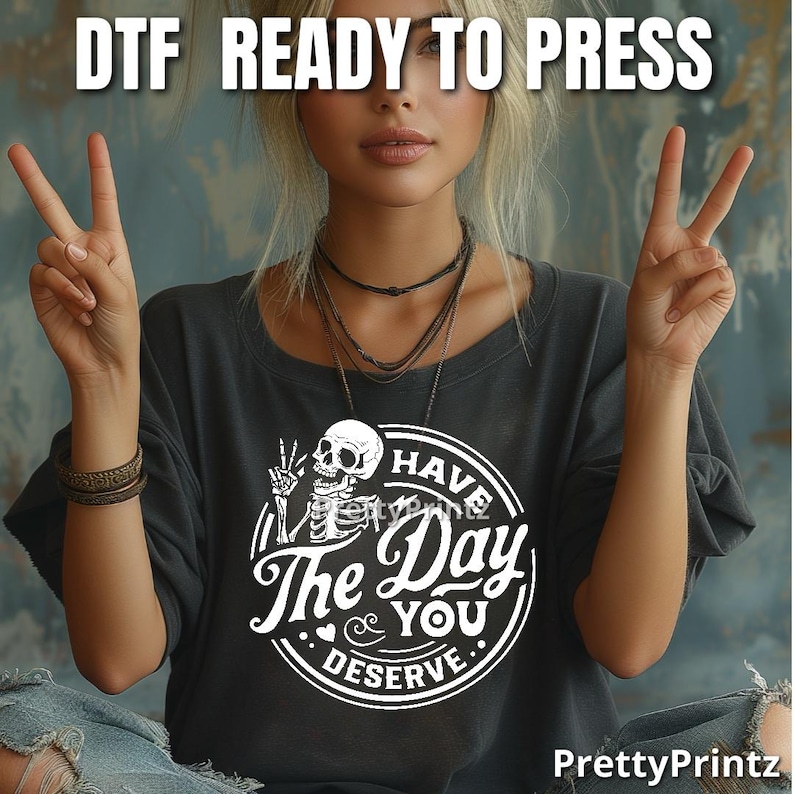 May include: A black t-shirt with a white graphic of a skeleton holding a peace sign and the text "Have the Day You Deserve".