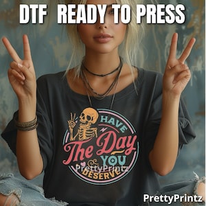 May include: A black t-shirt with a colorful graphic of a skeleton holding a peace sign and the text "Have The Day You Deserve".