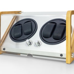 May include: A watch winder with a light wood frame and a clear acrylic cover. The device features two circular black rotating compartments, each containing two black textured watch pillows. Two silver control knobs are positioned on the front panel.