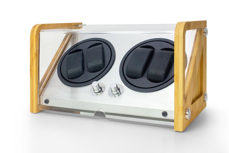 May include: A watch winder with a light wood frame and a clear acrylic cover. The device features two circular black rotating compartments, each containing two black textured watch pillows. Two silver control knobs are positioned on the front panel.