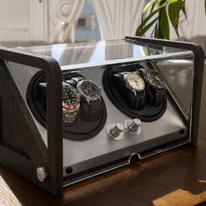 May include: A wooden watch winder with a clear acrylic top and stainless steel accents. The winder holds four watches, each displayed on a black cushion. The frame is dark brown, and the winder sits on a round wooden table.