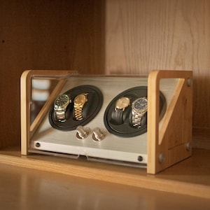 May include: A wooden watch winder with a clear acrylic cover, featuring two circular rotating slots that hold four wristwatches. The device has a light wood frame and a silver-toned interior panel with two control knobs on the front.