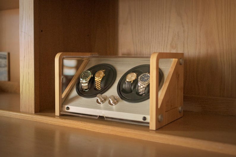 May include: A wooden watch winder with a clear acrylic cover, featuring two circular rotating slots that hold four wristwatches. The device has a light wood frame and a silver-toned interior panel with two control knobs on the front.