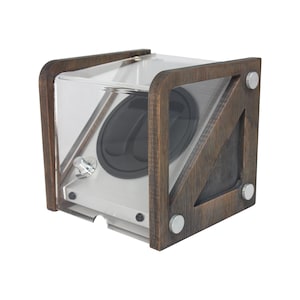 May include: A watch winder with a clear acrylic front and sides, set within a dark brown wooden frame. The winder has a silver-colored interior with a black circular component and silver hardware. The wooden frame is secured with silver-colored fasteners.