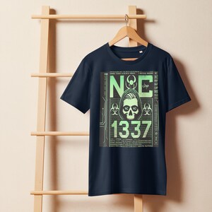 以下が含まれることがあります： 緑と黒のグラフィックプリントが施されたネイビーブルーのTシャツ。フードをかぶった人物と「N&C 1337」の文字が描かれています。