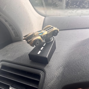 May include: A bronze-coloured toy car with blue windows mounted on a black rectangular base. The base features a white dashed line painted down the centre, resembling a road. The car has a detailed engine block visible on the rear deck.