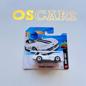 Puede incluir: Un coche de juguete blanco '92 Dodge Viper RT/10 fundido a presión sobre una tarjeta azul y blanca. La tarjeta presenta el texto "OSCARS" y "DROP TOPS". El coche de juguete tiene ruedas negras y detalles en azul. La tarjeta también tiene el texto "2/5".