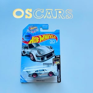 May include: A Hot Wheels die-cast toy car in a package. The car is silver with pink wheels and the number 34. The package has the Hot Wheels logo, the word "OSCARS", and the text "NIGHTBURNERZ".