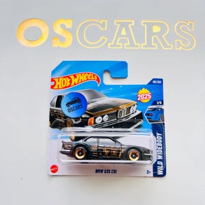 Puede incluir: Modelo a escala Hot Wheels de un BMW 635 CSI negro con ruedas y detalles dorados. El embalaje incluye el logotipo de Hot Wheels, el texto "OSCARS" y "WILD WIDEBODY". El modelo está marcado como 185/250 y 1/5.