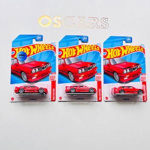 Può includere: Tre modellini di auto Hot Wheels rosse, confezionate su schede blu e rosse. Le schede presentano il logo Hot Wheels e una grafica dell'auto. Il testo "RED EDITION" è visibile. Le auto sono modelli BMW M3.