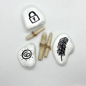 May include: White painted stones with black ink drawings of a lock, a feather, and a spiral sun symbol. Two rolled scrolls tied with twine are also present. The items are arranged on a white surface.