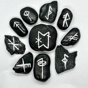 Könnte beinhalten: Zehn schwarze Steine mit weißen Runensymbolen, kreisförmig angeordnet. Die Steine haben unterschiedliche Formen und Größen. Die Runen sind in einem einfachen, kräftigen Stil gemalt.