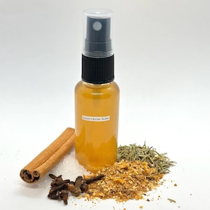 Könnte beinhalten: Eine transparente Glassprühflasche gefüllt mit bernsteinfarbener Flüssigkeit, beschriftet mit "Autumn Ember Water". Zimtstangen, Nelken, Salz und getrocknete Kräuter umgeben die Flasche. Sie hat eine schwarze und transparente Sprühdüse.