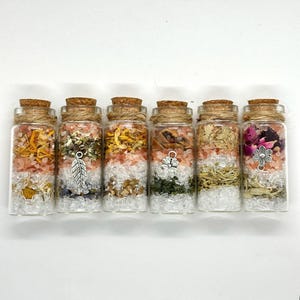 May include: Six small glass bottles with cork stoppers, filled with layers of colorful herbs, crystals, and salts. Each bottle is adorned with a silver charm and tied with twine. The contents include pink, white, yellow, and green elements.