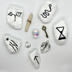 May include: A collection of mystical objects, including white painted stones with black symbols, a rolled scroll, a small key, a leaf charm, and a glass orb. The stones feature designs like an hourglass, a snake, and an arrow.