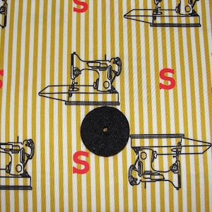 May include: A black circular object with a hole in the center is centered on a fabric with a yellow and white striped pattern. The fabric has a repeating pattern of black sewing machines and red letter "S"s.