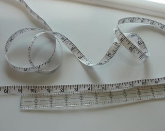 Puede incluir: Una cinta métrica blanca con marcas negras está enrollada sobre una superficie blanca. También se ve una regla transparente con marcas negras.