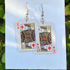Puede incluir: Un par de pendientes con la carta del Rey de Diamantes. Los pendientes están hechos de metal y tienen un acabado plateado. La carta de juego está impresa sobre un fondo blanco con detalles rojos y negros.