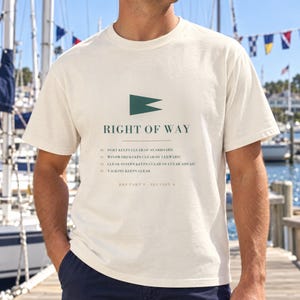 May include: A cream-colored short-sleeve t-shirt featuring a dark green nautical flag icon and the text RIGHT OF WAY. Below the text, it lists sailing rules: 10 Port keeps clear of starboard, 11 Windward keeps clear of leeward, 12 Clear astern keeps clear of clear ahead, 13 Tacking keeps clear, and RRS Part 2 Section A.