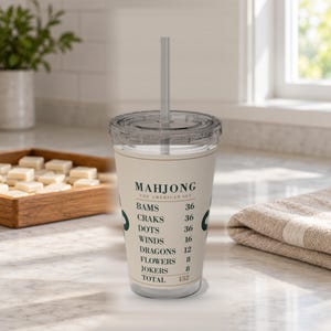 Puede incluir: Un vaso de pl&aacute;stico transparente con tapa y pajita, que presenta una banda de color crema impresa con texto verde oscuro que dice Mahjong The American Set, seguido de una lista de categor&iacute;as de juego y valores num&eacute;ricos que suman 152.