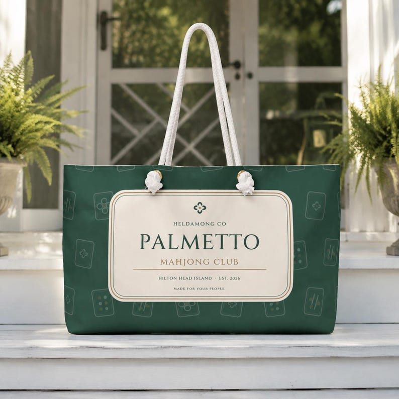 May include: A dark green tote bag with white rope handles and a rectangular cream-colored label. The label features the text HELDAMONG CO PALMETTO MAHJONG CLUB HILTON HEAD ISLAND EST. 2026 MADE FOR YOUR PEOPLE. The bag has a repeating white line art pattern.