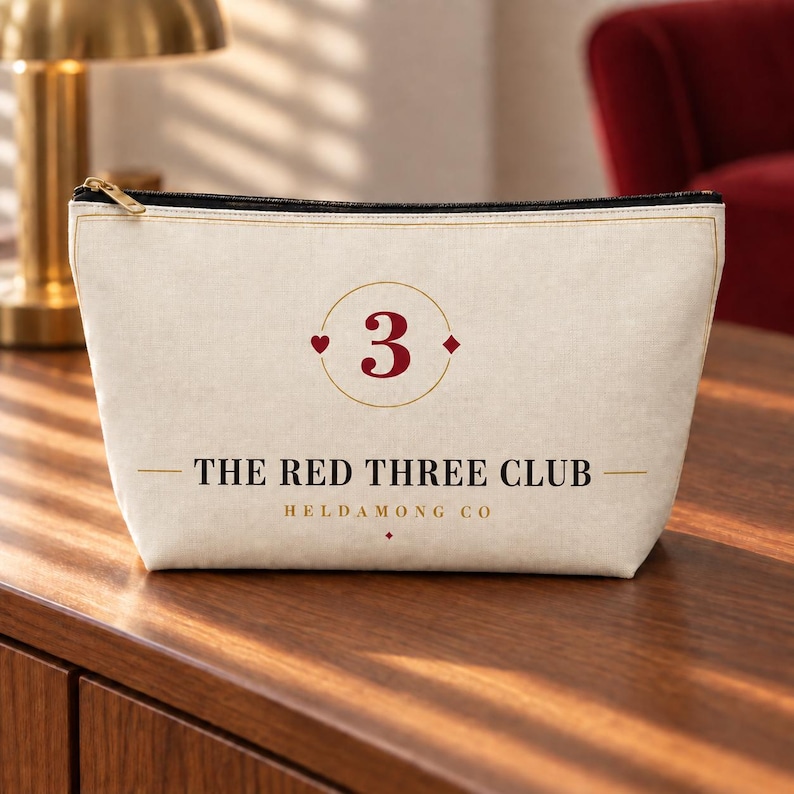 K&ouml;nnte beinhalten: Eine cremefarbene Rei&szlig;verschlusstasche aus Segeltuch mit einer zentralen Grafik, die die Zahl 3, ein rotes Herz und ein rotes Karo zeigt. Darunter steht der Text THE RED THREE CLUB und HELDAMONG CO in schwarzer und goldener Schrift.