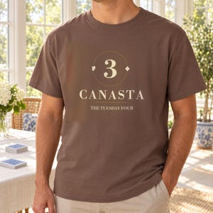 Puede incluir: Una camiseta marr&oacute;n de manga corta con un gr&aacute;fico que muestra el n&uacute;mero 3 dentro de un c&iacute;rculo, el texto CANASTA y el texto THE TUESDAY FOUR. El dise&ntilde;o incluye peque&ntilde;os s&iacute;mbolos de cartas como un coraz&oacute;n y un diamante junto al n&uacute;mero 3.
