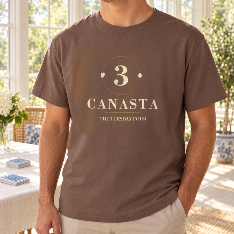 Puede incluir: Una camiseta marr&oacute;n de manga corta con un gr&aacute;fico que muestra el n&uacute;mero 3 dentro de un c&iacute;rculo, el texto CANASTA y el texto THE TUESDAY FOUR. El dise&ntilde;o incluye peque&ntilde;os s&iacute;mbolos de cartas como un coraz&oacute;n y un diamante junto al n&uacute;mero 3.