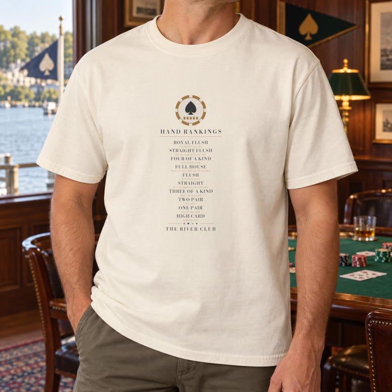 May include: A cream-colored short-sleeve t-shirt featuring a central graphic of a gold and black poker chip with a spade icon. Below the icon, the text reads HAND RANKINGS followed by a list of poker hands and the words THE RIVER CLUB.