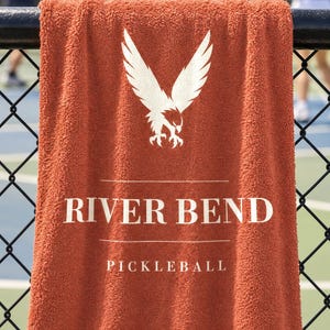 May include: A burnt orange terry cloth towel featuring a white graphic of an eagle with wings spread. The text reads RIVER BEND, PICKLEBALL, and HELDAMONG CO in white lettering. The towel is draped over a black chain-link fence.