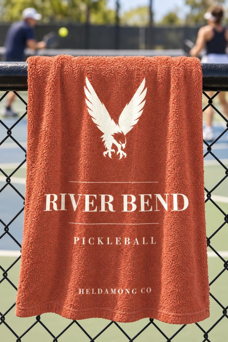 May include: A burnt orange terry cloth towel featuring a white graphic of an eagle with wings spread. The text reads RIVER BEND, PICKLEBALL, and HELDAMONG CO in white lettering. The towel is draped over a black chain-link fence.