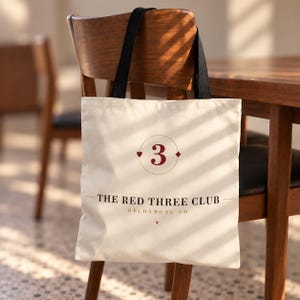 May include: A cream-coloured canvas tote bag with black shoulder straps. The front features a graphic with a large red number 3 inside a circle, flanked by a heart and diamond symbol. Below, the text reads THE RED THREE CLUB HELDAMONG CO.