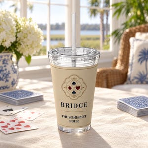 May include: A clear plastic tumbler with a lid and straw, featuring a beige wrap printed with a decorative emblem of four card suits, the word BRIDGE, and the text THE SOMERSET FOUR. The design includes a black spade, red diamond, red heart, and black club.