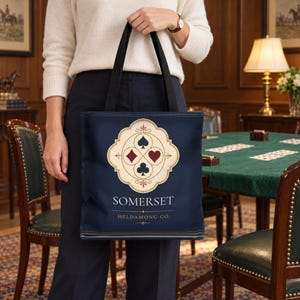 May include: A navy blue tote bag with black handles featuring a cream-coloured ornate emblem containing the four playing card suits: spade, diamond, heart, and club. The text SOMERSET and HELDAMONG CO. is printed in white below the emblem.
