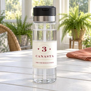 May include: A clear plastic water bottle with a black flip-top lid and a white straw inside. The bottle features a cream-colored label with the number 3, the word CANASTA, and the text THE THURSDAY FOUR printed in dark red with gold accents.