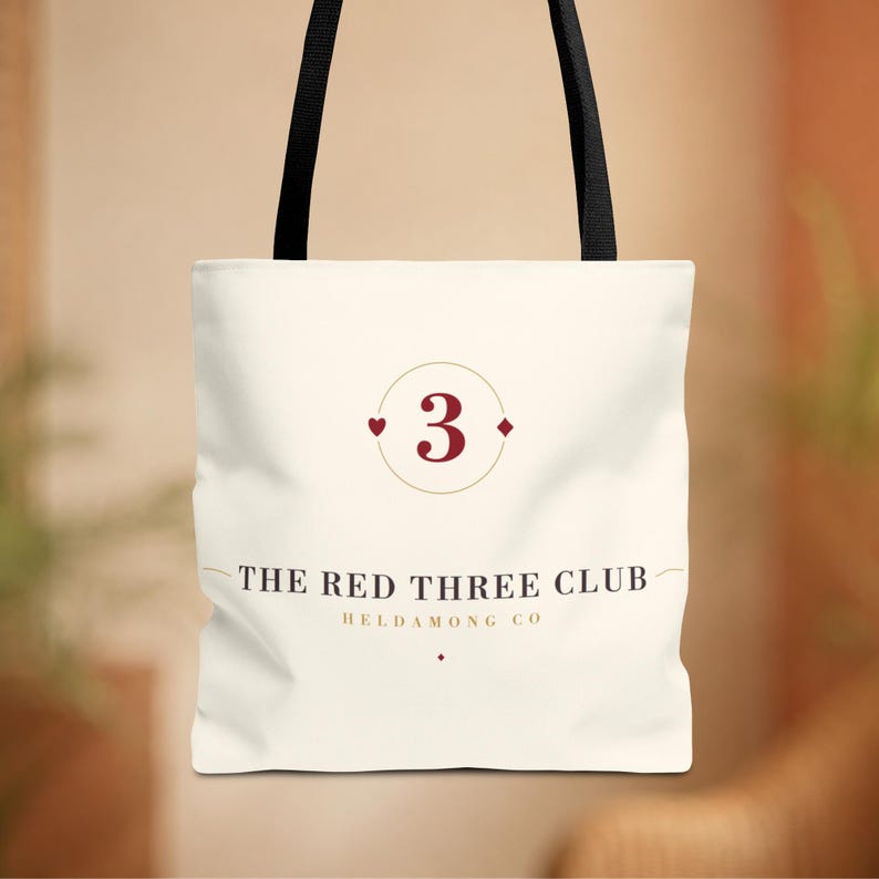 May include: A cream-coloured tote bag with black shoulder straps. The front features a graphic with a large red number 3 inside a circle, flanked by a small red heart and diamond. Below, the text reads THE RED THREE CLUB HELDAMONG CO.
