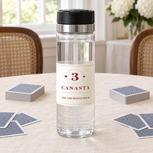 May include: A clear plastic water bottle with a black lid and a white straw. The bottle features a cream-colored label with the number 3, the word CANASTA, and the text THE THURSDAY FOUR printed in dark red, surrounded by small red card suit symbols.