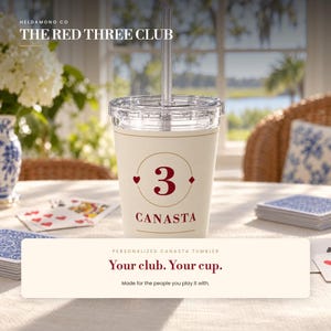 May include: A cream-colored tumbler with a clear lid and straw, featuring a red number 3 inside a gold circle, flanked by a heart and diamond symbol, with the word CANASTA printed below in red. The design is centered on the front of the cup.