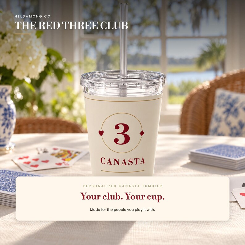 May include: A cream-colored tumbler with a clear lid and straw, featuring a red number 3 inside a gold circle, flanked by a heart and diamond symbol, with the word CANASTA printed below in red. The design is centered on the front of the cup.