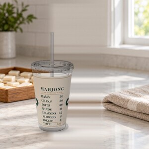 Puede incluir: Un vaso de pl&aacute;stico transparente con tapa y pajita, que presenta una banda de color crema impresa con texto verde oscuro que dice MAHJONG THE AMERICAN SET, seguido de una lista de categor&iacute;as de juego y sus valores num&eacute;ricos que suman 152.