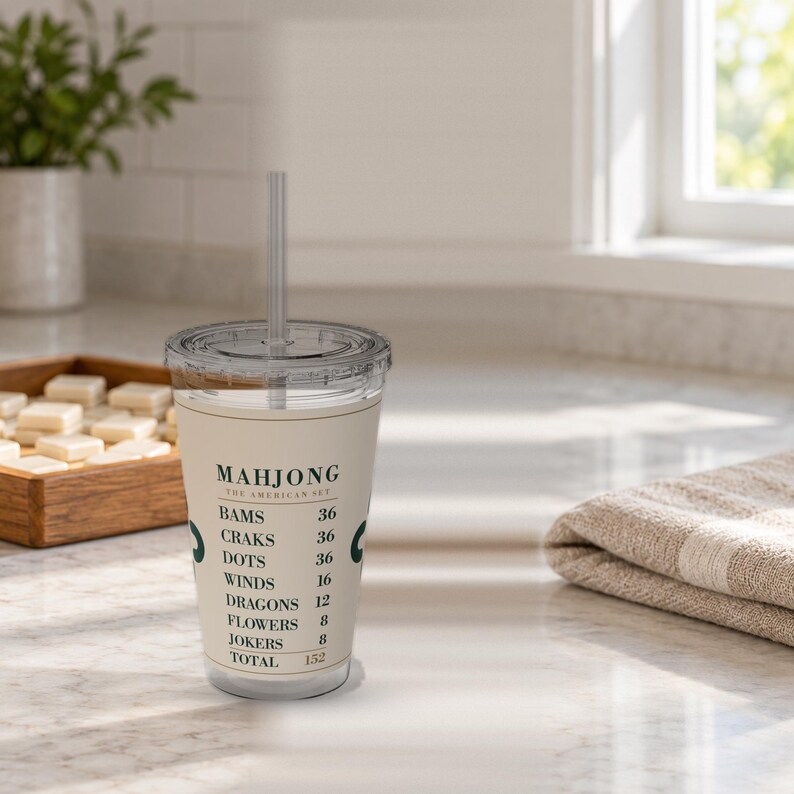 Puede incluir: Un vaso de pl&aacute;stico transparente con tapa y pajita, que presenta una banda de color crema impresa con texto verde oscuro que dice MAHJONG THE AMERICAN SET, seguido de una lista de categor&iacute;as de juego y sus valores num&eacute;ricos que suman 152.