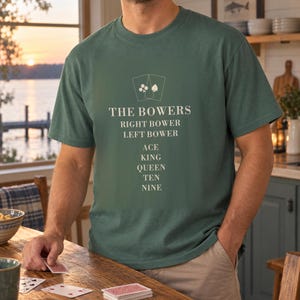 May include: A dark green short-sleeve t-shirt featuring a cream-colored graphic of two playing cards above the text THE BOWERS, RIGHT BOWER, LEFT BOWER, ACE, KING, QUEEN, TEN, NINE printed in a stacked, centered, sans-serif font on the chest.