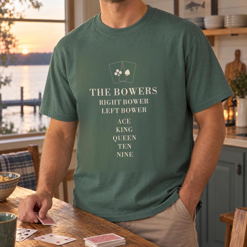 May include: A dark green short-sleeve t-shirt featuring a cream-colored graphic of two playing cards above the text THE BOWERS, RIGHT BOWER, LEFT BOWER, ACE, KING, QUEEN, TEN, NINE printed in a stacked, centered, sans-serif font on the chest.