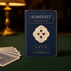 Puede incluir: Una baraja de cartas rectangular en una caja azul oscuro con letras doradas. La caja dice SOMERSET BRIDGE CLUB y HELDAMONG CO. Un emblema central de color crema muestra los cuatro palos de la baraja en rojo y azul, con iconos m&aacute;s peque&ntilde;os.